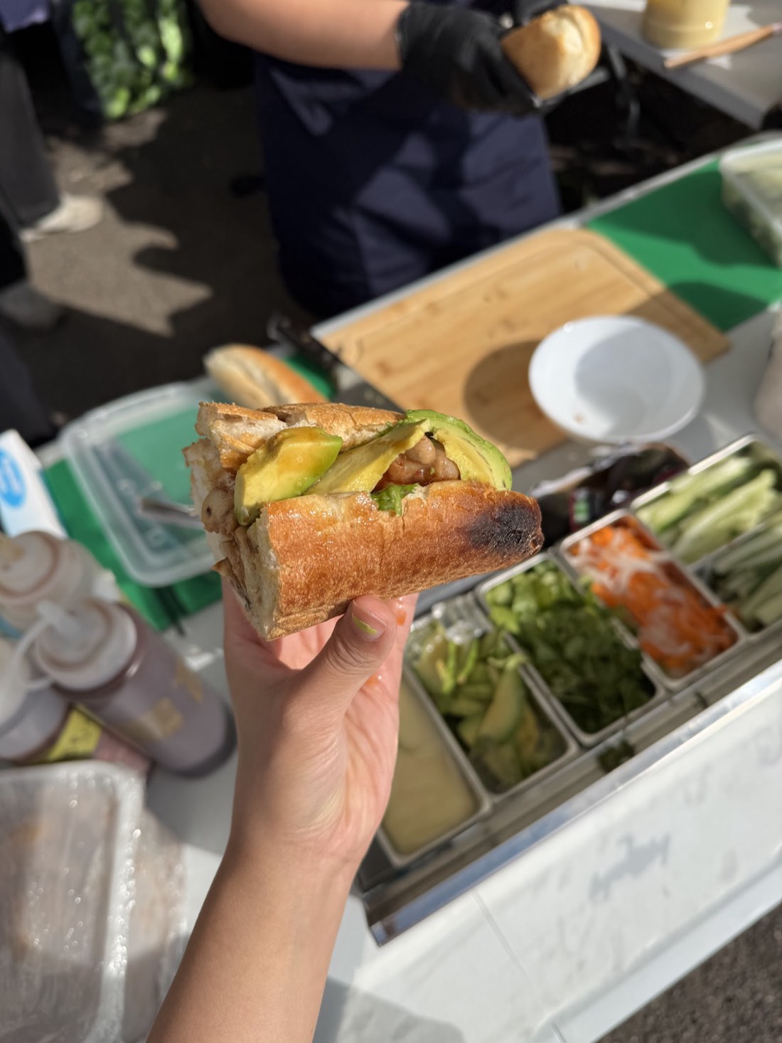 A Honey Chilli Chicken Avolcano bánh mì held up in one hand, avocado and chicken visible through the open baguette, with the NgON prep station and trays of pickled carrot, coriander, cucumber and avocado behind it.
