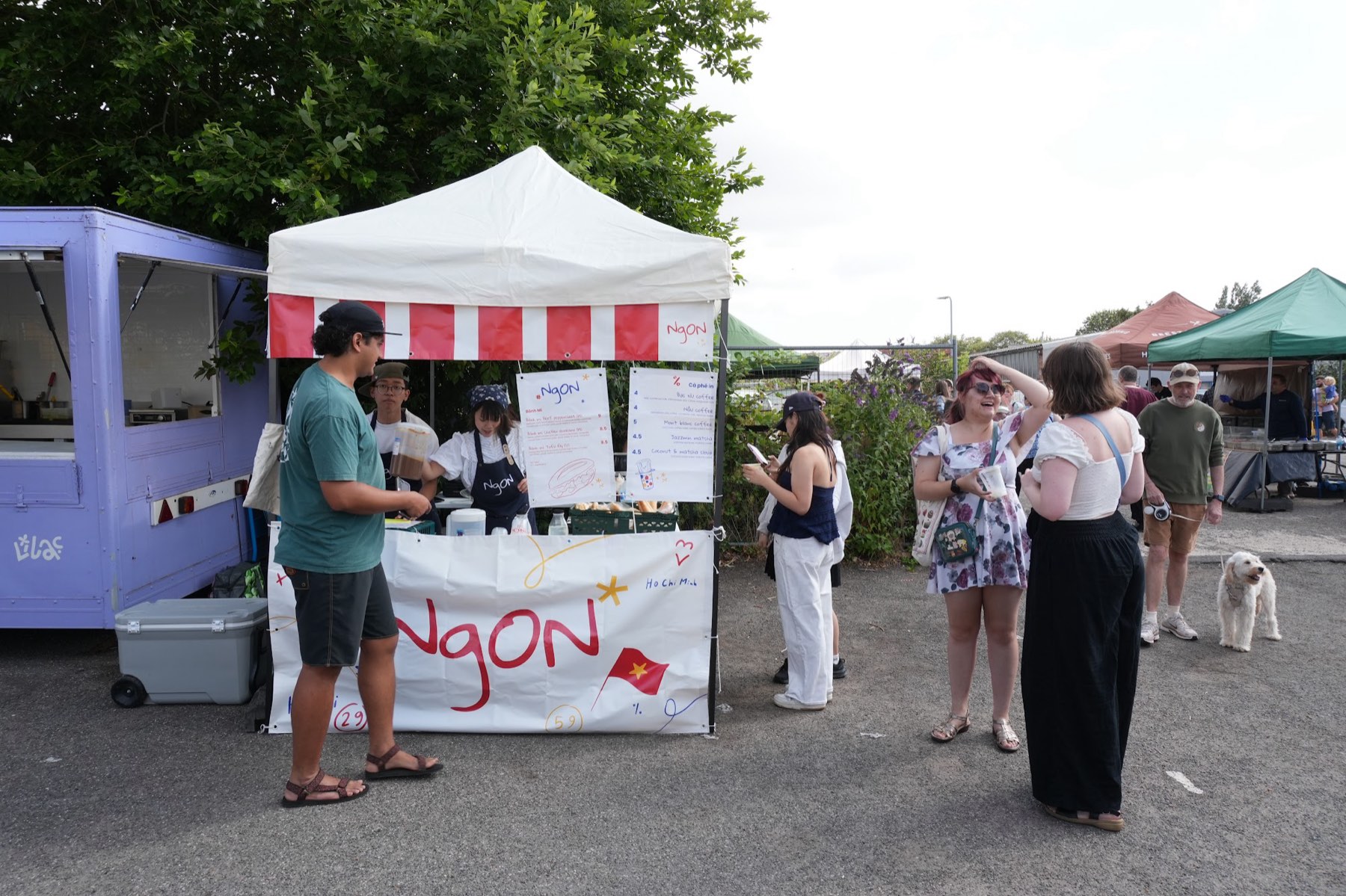 NgON's white gazebo with a red-and-white striped valance at The Garage, Chilwell. The handwritten NgON logo runs across a counter banner with hand-drawn artwork and a Vietnamese flag, two menu boards hang above, team members in NgON aprons work behind the counter and customers stand nearby holding cups.
