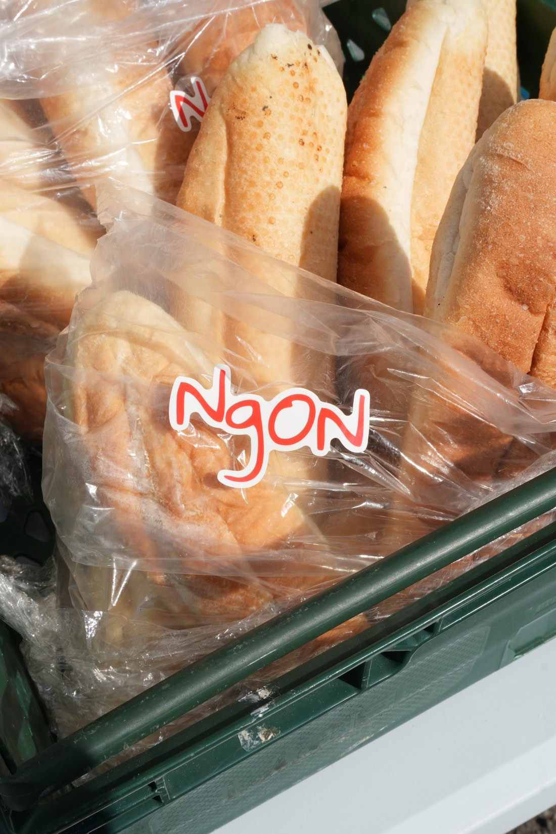 Bánh mì baguettes in a green crate, each bag sealed with a red handwritten NgON sticker, lit by direct sunlight.