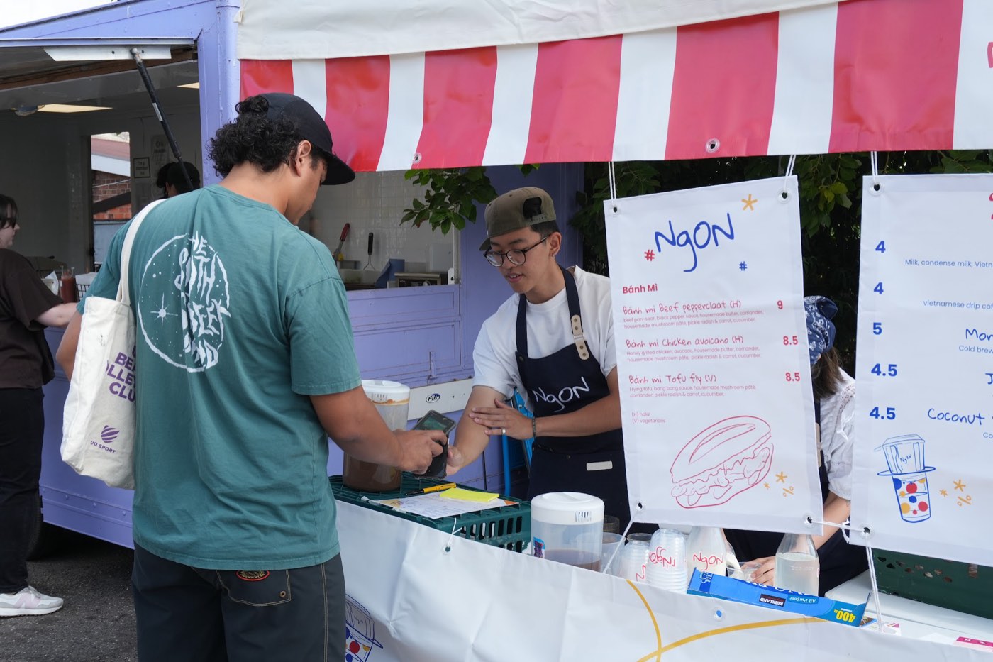 A customer paying by card at the NgON counter while a team member in a NgON apron takes the order. The bánh mì menu board shows Beef Pepperclaat, Chicken Avolcano and Tofu Fly with halal and vegetarian markings, and branded NgON cups sit on the counter.