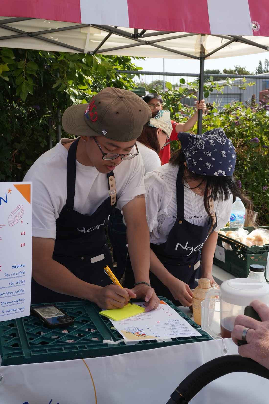 Two NgON team members in navy aprons leaning over the counter, one writing an order on a yellow sticky note. The cà phê menu board lists Bạc xỉu, Nâu, Mont blanc, Jazzmin matcha and Coconut and matcha cloud, with a crate of bagged baguettes behind them.