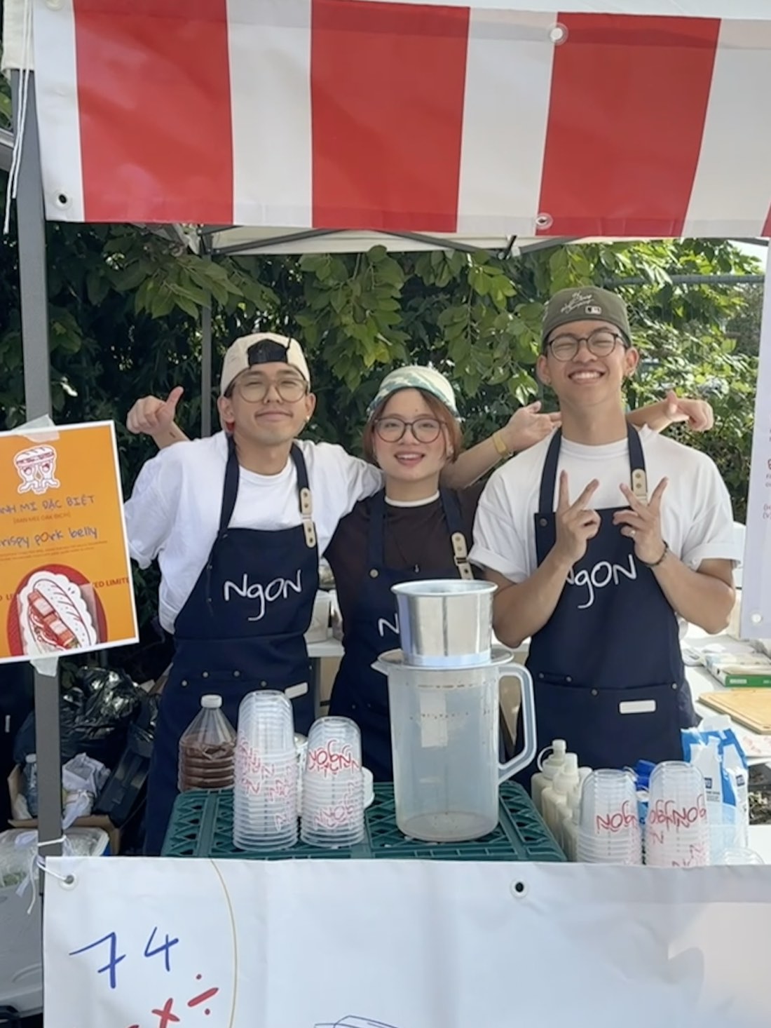 Three of the NgON team in navy NgON aprons behind the counter under the red-and-white striped gazebo, arms around each other. Stacked NgON cups and a Vietnamese phin coffee filter sit on the counter in front of them.